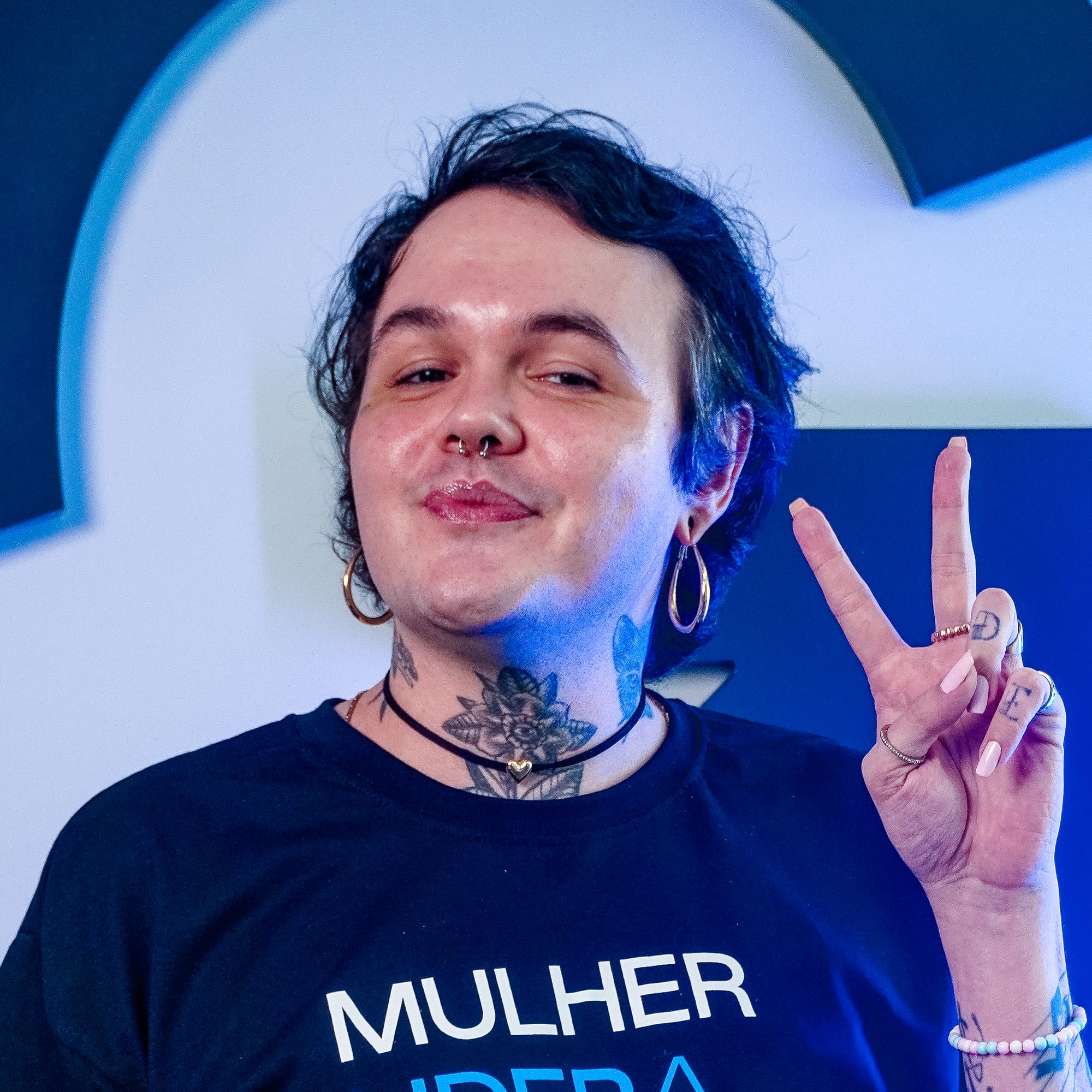 Pessoa com cabelo curto e camiseta preta com a frase 'Mulher lidera onde ela quiser', fazendo sinal de paz com os dedos.