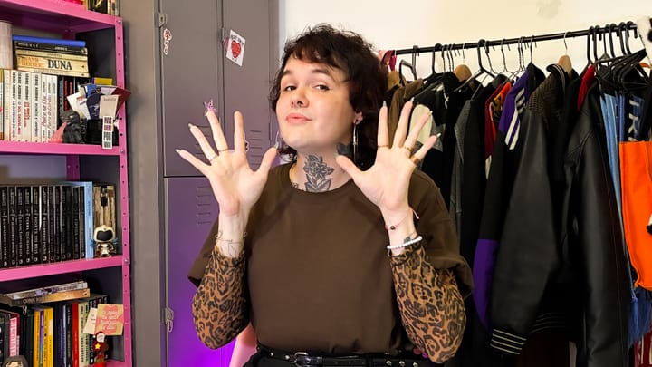 Mulher trans branca e tatuada fazendo o número 10 com as mãos.